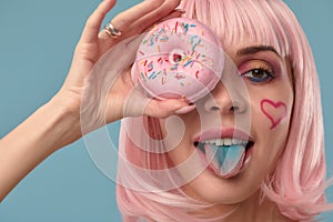 Portrait of a fashionable girl in a pink wig and with a pink donut in her hands keeps to the eye, shows tongue
