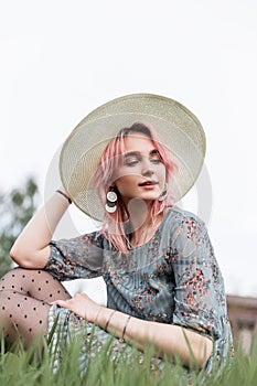 Portrait fashionable pretty young woman model with chic pink hair in hat in stylish blue dress with floral print on grass in field
