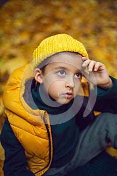 portrait of a fashionable serious child boy autumn sitting on a trail in orange leaves in the afternoon at a green fence