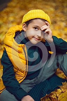 portrait of a fashionable serious child boy autumn sitting on a trail in orange leaves in the afternoon at a green fence