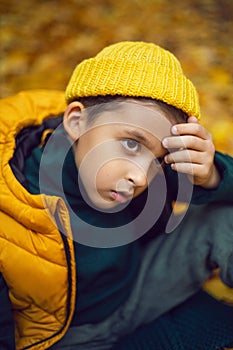 portrait of a fashionable serious child boy autumn sitting on a trail in orange leaves in the afternoon at a green fence