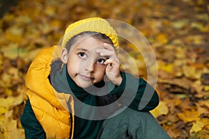 portrait of a fashionable serious child boy autumn sitting on a trail in orange leaves in the afternoon at a green fence