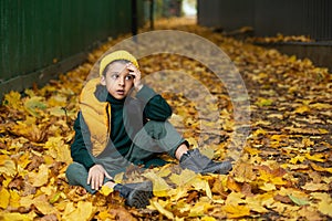 portrait of a fashionable serious child boy autumn sitting on a trail in orange leaves in the afternoon at a green fence