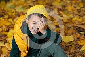 portrait of a fashionable serious child boy autumn sitting on a trail in orange leaves in the afternoon at a green fence