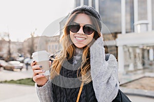 Portrait fashionable urban young woman with coffee to go walking in sunny city centre. Amazing smiling girl in modern