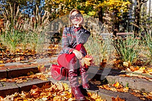 Portrait of fashionable woman wearing stylish blazer, skirt, knee high leather boots sitting in fall park with purse