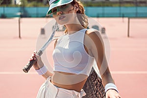 portrait of fashionable woman in white clothing and cap with tennis racket posing at tennis net
