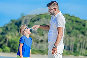 Portrait of father giving water to his thirsty handsome boy on the desert tropical beach.