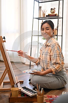 Portrait of female artist holding palette and painting picture on canvas with oil paints in cozy home studio