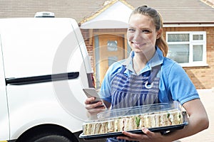 Portrait Of Female Caterer Using Mobile Phone Delivering Tray Of Sandwiches To House