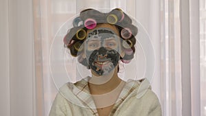 Portrait of female with curlers on head with clay cleansing black mask on face, closeup. Young woman taking care of skin complexio