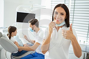 Portrait of female dentist showing thumb up while her collegue working with small client on the background
