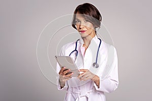 Portrait of a female doctor holding her patient chart on digital tablet while standing against light grey background