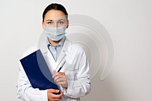 Portrait of a female doctor in a protective mask with a clipboard and a pen in her hands, making a record