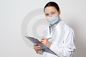Portrait of a female doctor in a protective mask with a clipboard and a pen in her hands, making a record