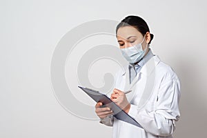 Portrait of a female doctor in a protective mask with a clipboard and a pen in her hands, making a record