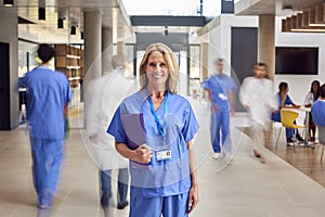 Portrait Of Female Doctor  Wearing Scrubs Holding Patient Notes In Busy Hospital
