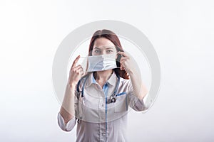 Portrait of a female doctor in a white uniform who puts on a medical mask in the studio on a white background.