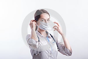 Portrait of a female doctor in a white uniform who puts on a medical mask in the studio on a white background.