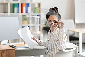 Portrait of female entrepreneur working with documents at desk in office, taking off glasses and smiling at camera