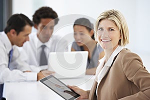 Portrait Of Female Executive Using Tablet Computer With Office Meeting In Background