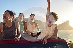 Portrait Of Female Friends Standing Up Through Sun Roof Car And Dancing On Road Trip 