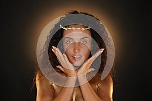 Portrait of female model in studio. Attractive girl wearing tiara and bright makeup covered in golden paint holding face