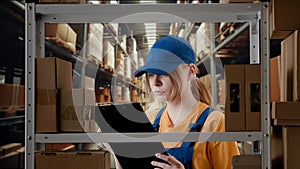 Portrait of female model working in storage. Girl storekeeper in uniform standing near rack with boxes, checking goods