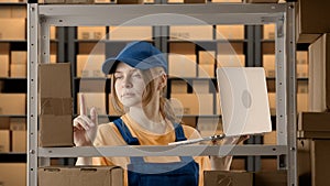 Portrait of female model working in storage. Girl storekeeper in uniform standing near rack with boxes, checking goods