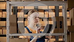 Portrait of female model working in storage. Girl storekeeper in uniform standing near rack with boxes, checking goods