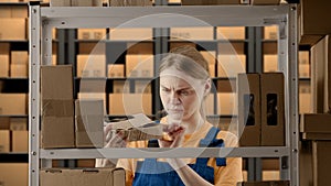 Portrait of female model working in storage. Girl storekeeper in uniform standing near rack with boxes finds damaged