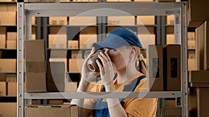 Portrait of female model working in storage. Girl storekeeper in uniform standing near rack with boxes, having break