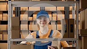 Portrait of female model working in storage. Girl storekeeper in uniform standing near rack with boxes, takes package