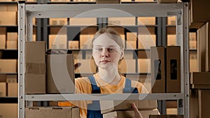 Portrait of female model working in storage. Girl storekeeper in uniform standing near rack with boxes, taking packages