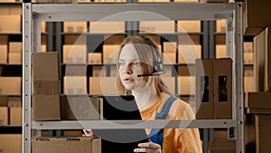 Portrait of female model working in storage. Girl storekeeper in uniform standing near rack with boxes, talking in