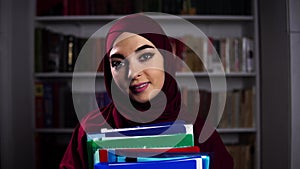 Portrait of a female Muslim secretary who stands on a bookshelf background with papers in her arms