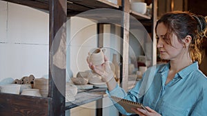 Portrait of female potter checking ceramics and writing information in notebook in workshop