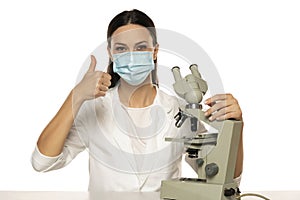 Portrait of female scientist with microscope and face mask showing thimbs up on a white background