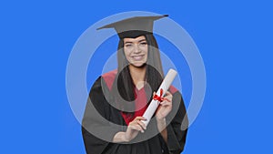 Portrait of female student in cap and gown graduation costume, holding diploma and showing ok gesture. Young woman