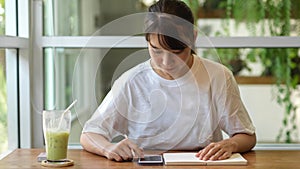 Portrait of female teenager using smartphone and writing an idea on a notebook in cafe
