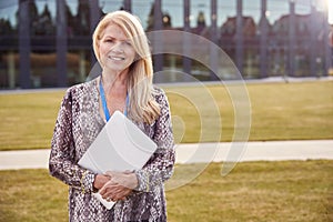 Portrait Of Female University Or College Tutor Outdoors With Modern Campus Building In Background