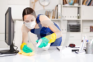 Portrait of a female worker in a protective mask, cleaning an office desk