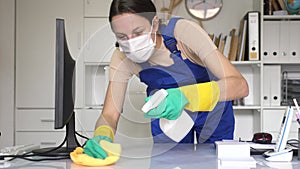 Portrait of a female worker in a protective mask, cleaning an office desk with a rag and detergent.