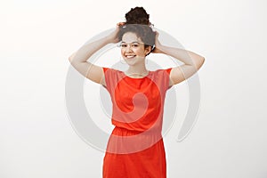 Portrait of femenine confident cute woman in stylish dress, touching combed hair and smiling with tender shy expression