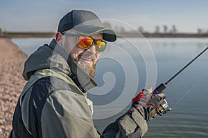 Portrait of fisherman. Bearded man in cap and sunglasses holds fishing rod at lake