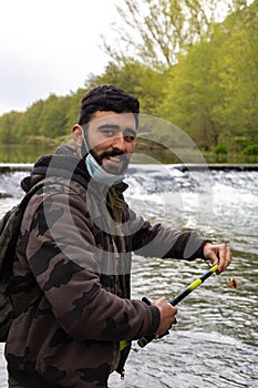 Portrait of a fisherman with camouflage suit, beard and mask by the covid19 smiling at the river bank holding the rod in his hand