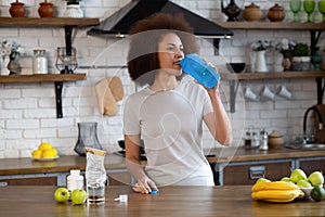 Portrait of fit black woman in sportswear at the kitchen is drinking from shaker with whey protein