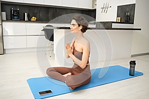 Portrait of fitness girl in kitchen, sits on yoga mat, meditates, listens meditation music in headphones, uses workout
