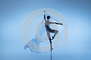 Portrait of flexible woman, graceful ballerina dancing with fabric, cloth isolated on blue studio background. Grace, art