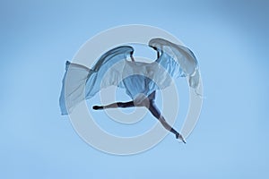 Portrait of flexible woman, graceful ballerina dancing with fabric, cloth isolated on blue studio background. Grace, art